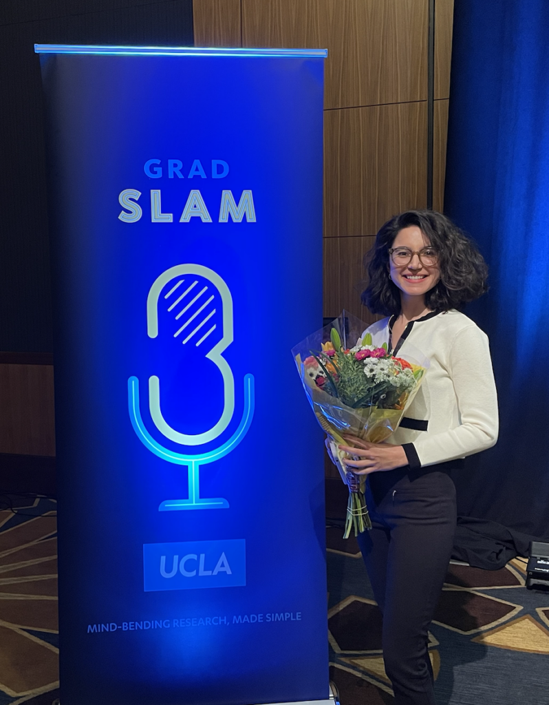 This is an image of graduate student Melis Cakar, holding a bouquet of flowers in front of a large blue sign for the 2024 UCLA Grand Slam.